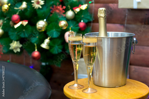 A metal bucket with a bottle of champagne in ice and two full glasses of sparkling wine on the background of a decorated Christmas tree.