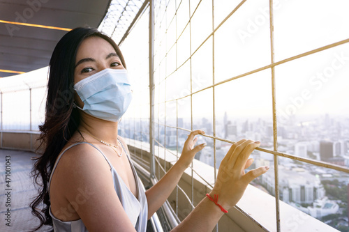 Young Asian beautiful woman with bronze dress wearing a hygiene protective mask over her face. Smiling female standing outside the building on the rooftop floor restaurant. New normal concept.