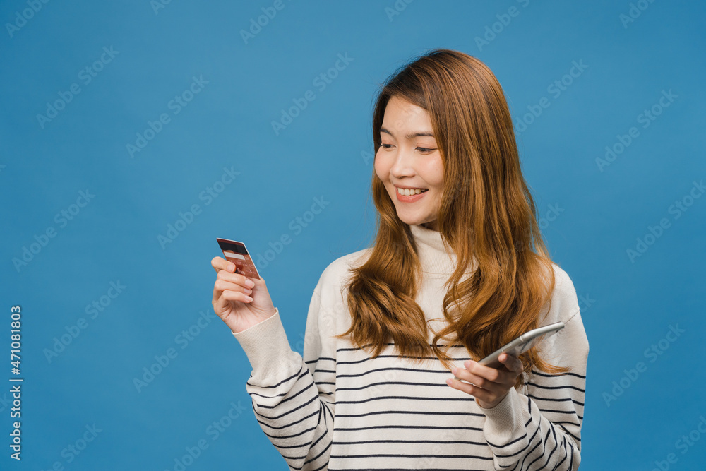 Young Asia lady using phone and credit bank card with positive expression, smiles broadly, dressed in casual clothing and stand isolated on blue background. Happy adorable glad woman rejoices success. Young Asia lady using phone and credit bank card with positive expression, smiles broadly, dressed in casual clothing and stand isolated on blue background. Happy adorable glad woman rejoices success.