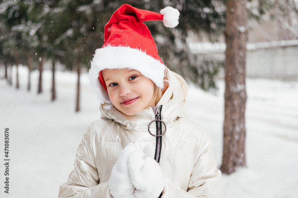 Obraz premium Laughing little girl in red christmas hat standing under snowfall looking at camera. Outdoors winter activities. Happy child in Santa hat playing, having fun in winter forest.