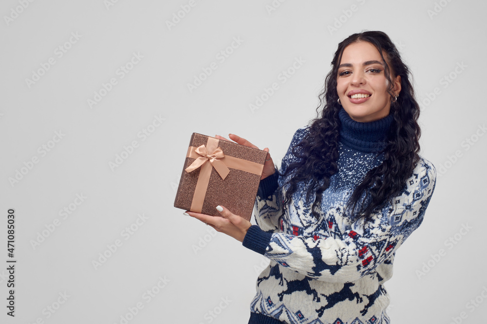 Pretty woman shows a gift box looking into the camera on a gray background, in a Christmas sweater. Pretty woman shows a gift box looking into the camera on a gray background, in a Christmas sweater.