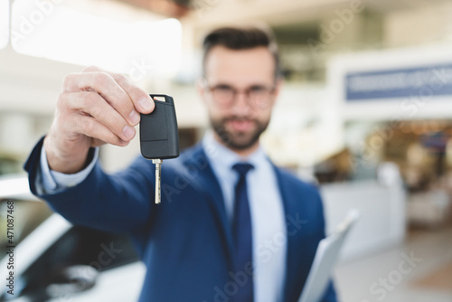 Focused smiling confident male shop assistant in formal clothes holding handing car keys to a client after buying purchasing new expensive car at automobile dealer shop. © InsideCreativeHouse