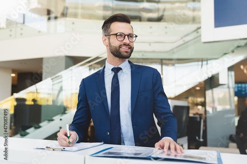 Successful caucasian smiling man shop assistant receptionist in formal attire writing while standing at reception desk in hotel car dealer shop photo