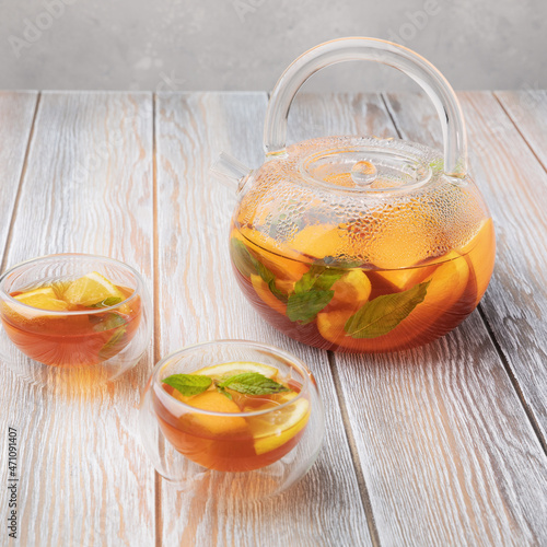 Fruit tea with orange, lemon and mint in glass teapot and two cups (bowls) on wooden background