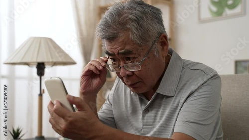 Elderly asian male having eyesight problems and wearing eyeglasses while gazing and reading message on smartphone on sofa. Old man having eye blurred vision indoors and moving glasses up and down 
