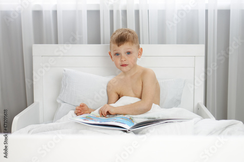Little boy with blue eyes is reading a book in a white bed