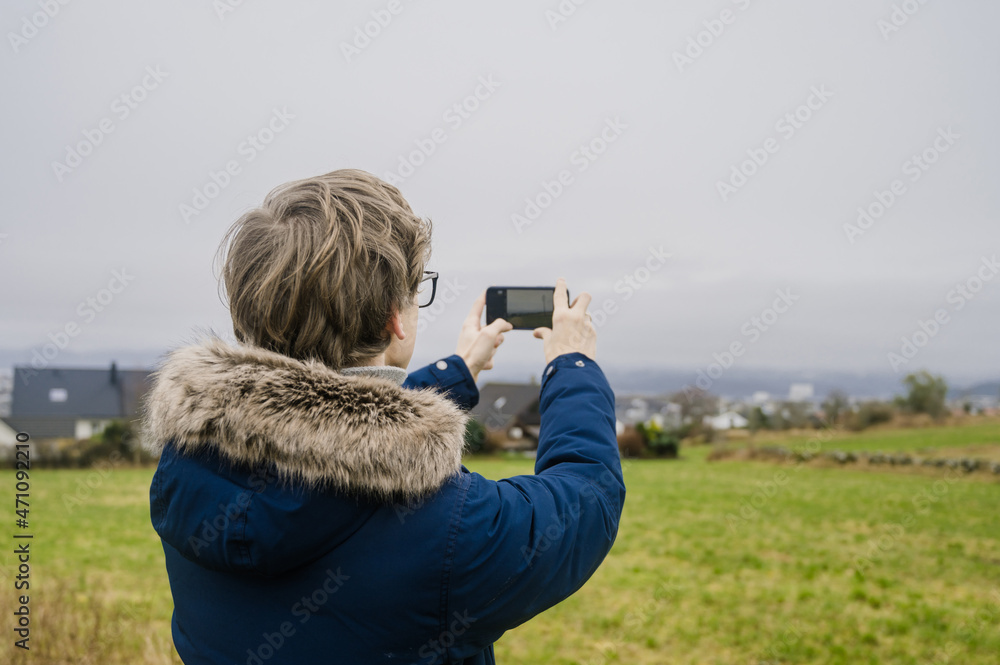 Obraz premium Young man in blue coat is taking a picture of city and green field by his phone. Cloudy day in Norway.