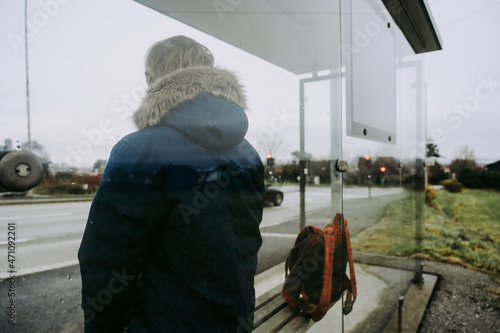 Man in blue coat with backpack is waiting for a bus. Modern glass bus stop on Norway. Cloudy day.