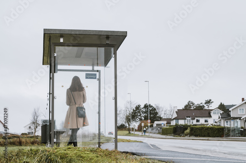 Elegant woman in coat is waiting for a bus. Modern glass bus stop on Norway. Cloudy day.