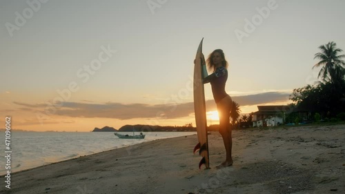 Young attractive female surfer smiling and posing with her surfboard at sunset on the beach