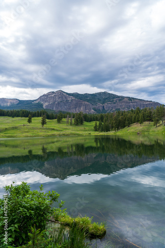Trout Lake Trail in Yellowstone National Park on a cloudy summer day - a mountain reflects in a blue lake, with green grass on the hillside in the distance