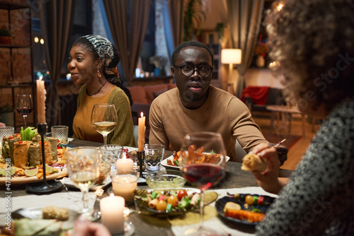 African young man in eyeglasses talking to his friends during dinner at the table