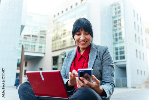 Hispanic business woman smiles while checking her mobile phone on the street.