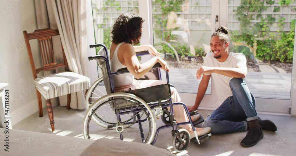 Happy biracial woman in wheelchair and smiling male partner talking in sunny living room Happy biracial woman in wheelchair and smiling male partner talking in sunny living room