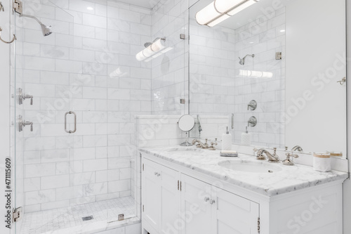 Fototapeta Naklejka Na Ścianę i Meble -  A luxurious white bathroom with a double vanity and bronze faucets, a white subway tile shower, and glass door.