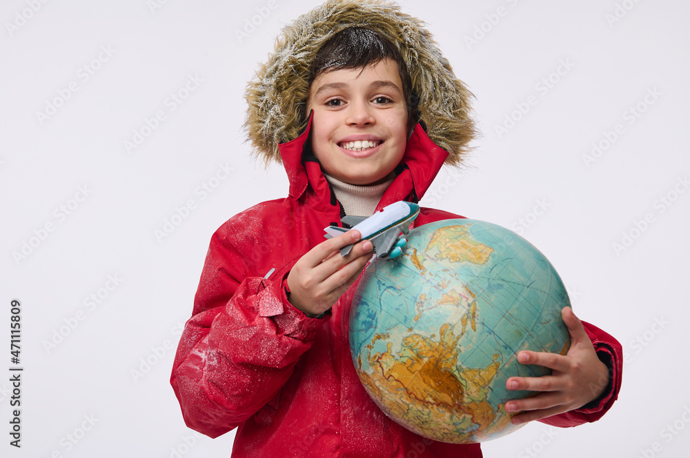 Around the world travel and winter tourism concept. Adorable elementary aged boy in warm bright red down jacket with hood holding an airplane toy model and globe, imitating the flight around the Earth Around the world travel and winter tourism concept. Adorable elementary aged boy in warm bright red down jacket with hood holding an airplane toy model and globe, imitating the flight around the Earth