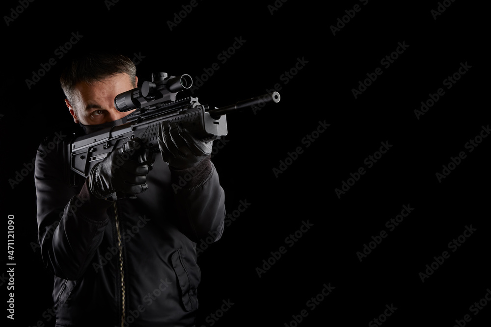 A man in dark clothes holds an automatic carbine in his hands. The face is covered with a mask, hands are in gloves. A figure with a weapon on a dark back A man in dark clothes holds an automatic carbine in his hands. The face is covered with a mask, hands are in gloves. A figure with a weapon on a dark back
