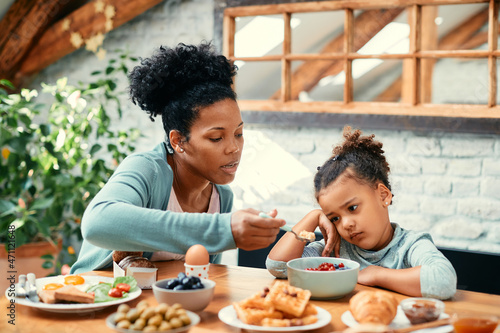 African American mother feeds her small daughter who is refusing to eat her breakfast at dining table.