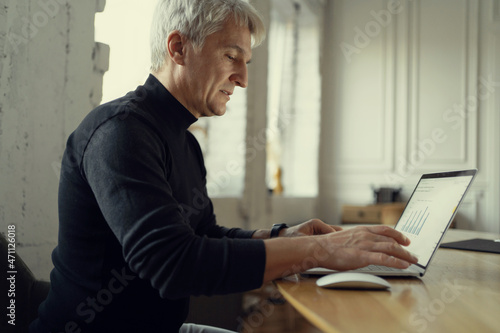 The designer creates projects online. An aged man works in a workplace. Prints a message to the client on the laptop. Communicates on a social network with colleagues.