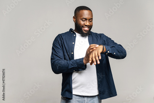 Positive African-American guy checking notification on the screen of smart watch, standing isolated on gray, smiling black man in casual wear looks at smart band on his wrist, controls time or rate photo