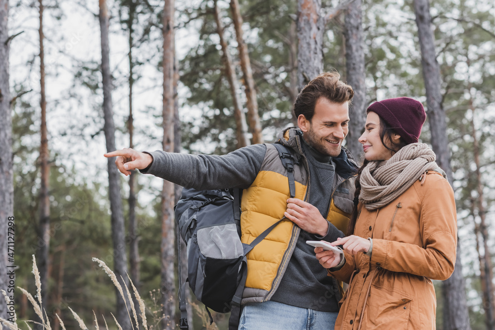 smiling man with backpack pointing with finger near girlfriend using smartphone in forest