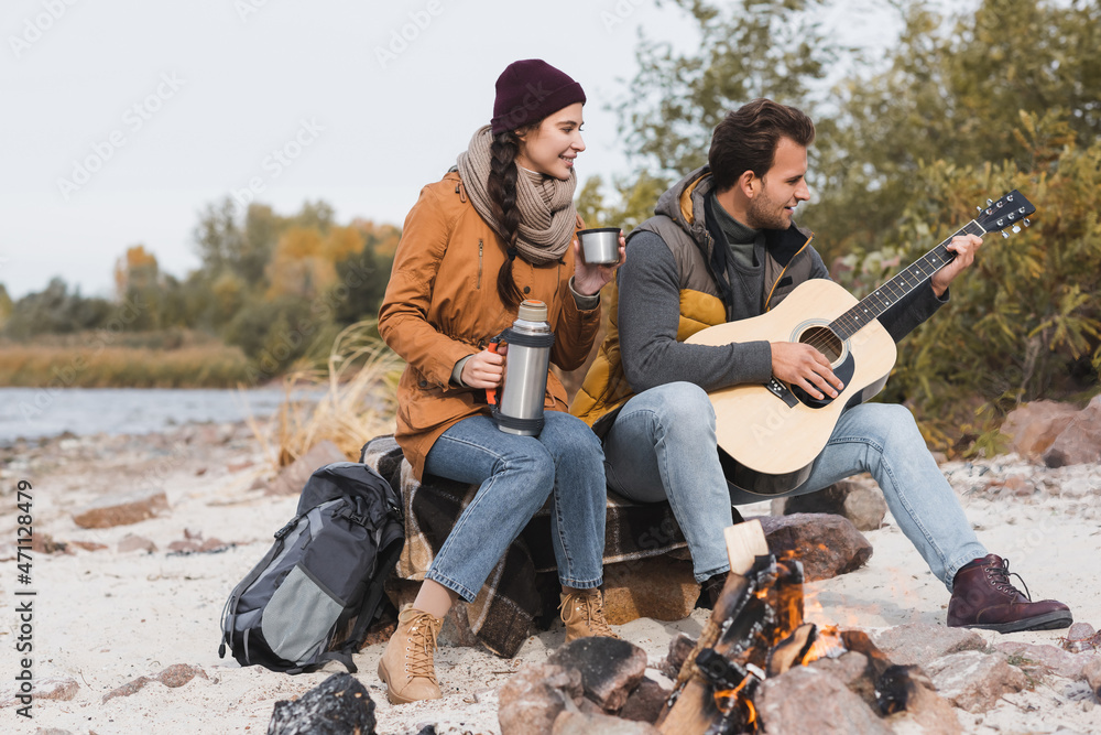woman with thermos and man with acoustic guitar sitting near bonfire woman with thermos and man with acoustic guitar sitting near bonfire