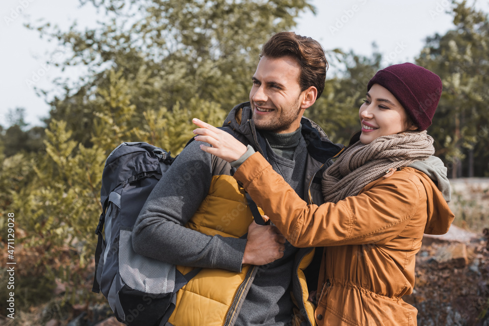 smiling woman pointing with finger near man with backpack while walking outdoors