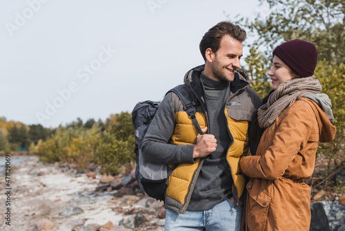 young woman with backpack smiling near girlfriend in autumn outfit while walking outdoors