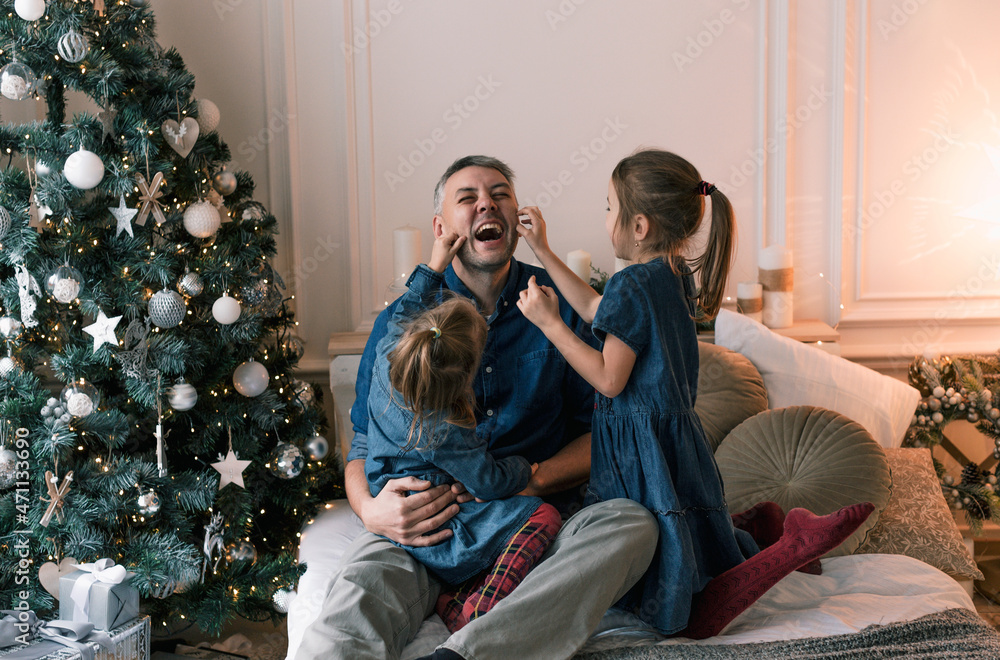 Dad and little daughters have fun together, sitting on the bed surrounded by pillows near the Christmas tree. happy christmas