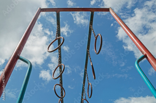 Photography Street monkey bars are metal sports rings hanging on chains in pairs at regular intervals on two crossbars attached parallel to racks, painted in blue and red, against background of cloudy sky