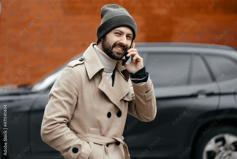 Φωτογραφία image of a young, smiling, happy, laughing man with a beard in a raincoat with a phone on a gray background on the street
