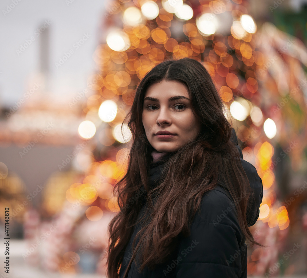 A beautiful and young girl looks into the camera. Christmas city decoration blurred in the background. Cute brunette faced to the camera in the downtown before New Year party