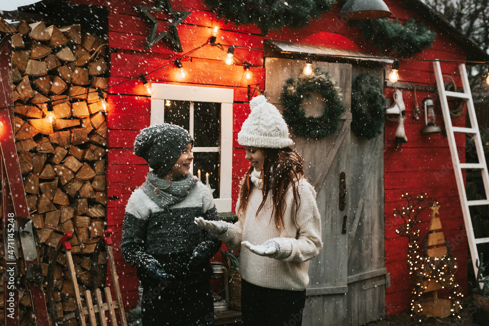 Obraz premium siblings teenage girl sister and cute boy brother in knitted sweater and hat having fun with first snow at porch of country house, concept of winter sports and Christmas holidays for children outdoor