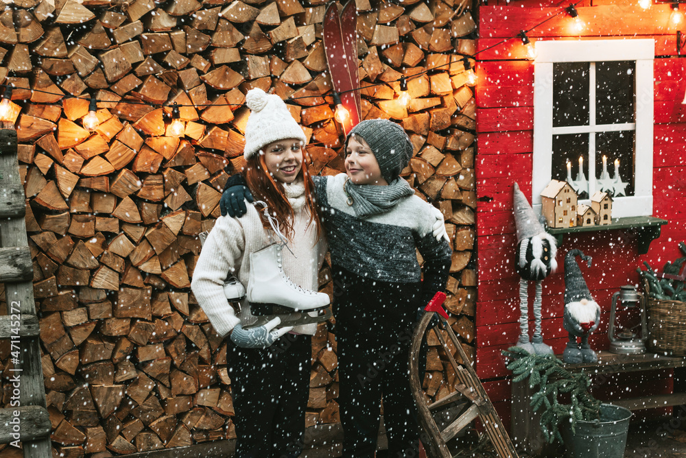 Obraz premium siblings teenage girl sister and cute boy brother in knitted sweater and hat stand at porch of country house with sleds and skates, concept of winter sports and Christmas holidays for children outdoor