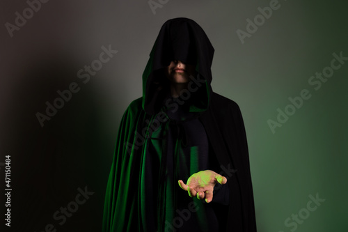 A mystical character in a black hoodie stretches out his hand to you. Witch in a black hood on her face, dark background green backlight