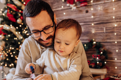 Father holding and playing with his son in living room decorated by Christmas tree and present gift box, the light give cozy atmosphere. New Year and merry christmas dad and baby concept