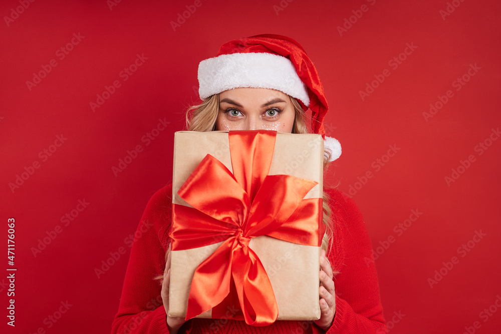 cheerful blonde in a Santa hat hid behind a gift box. Merry Christmas. Isolate on red background cheerful blonde in a Santa hat hid behind a gift box. Merry Christmas. Isolate on red background