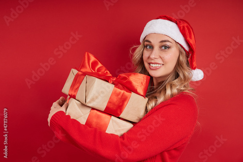 Charming smiling blonde in a Santa hat in a red sweater holding Christmas gifts. Isolate on red background