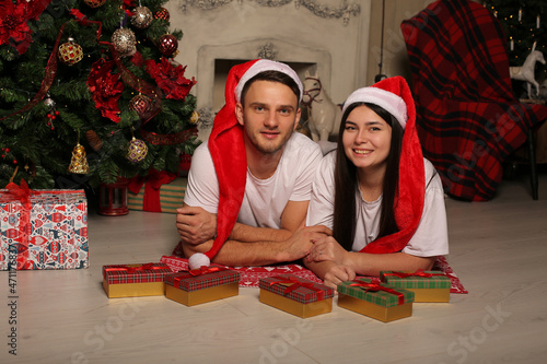 a young couple a guy and a girl in white T-shirts in a Santa Claus hat are lying on the floor near the Christmas tree next to gift boxes