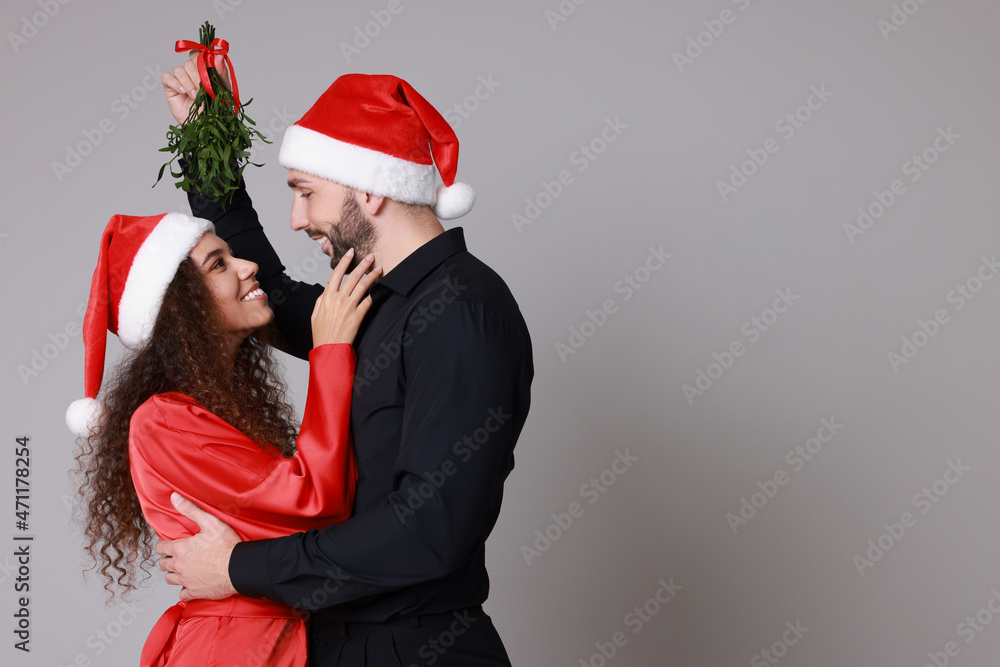 Obraz premium Lovely couple in Santa hats under mistletoe bunch on grey background. Space for text