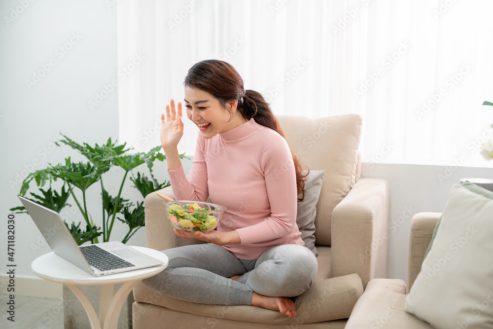 Beautiful young woman eating a healthy salad in living room, using a laptop computer, interior Beautiful young woman eating a healthy salad in living room, using a laptop computer, interior