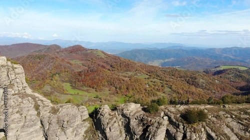 Aerial: two hikers walking along a very narrow mountain crest in autumn season. Forest with trees in yellowish colours, surrounding mountains and a valley photo