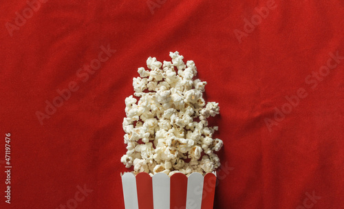 Popcorn scattered from a paper striped box on a red carpet. Traditional cardboard box or bucket with spilled kernels. Cinema film or movie theater composition.