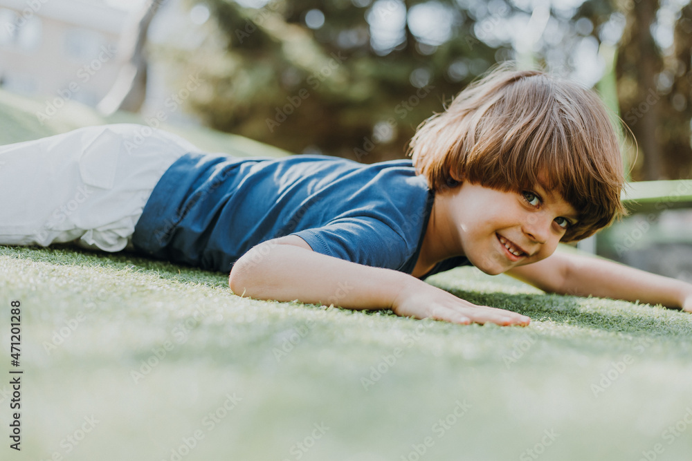little brunette boy in a blue T-shirt and white shorts is doing push-ups on a street sports field. child plays and trains outdoors. lifestyle. products for sports and health. space for text little brunette boy in a blue T-shirt and white shorts is doing push-ups on a street sports field. child plays and trains outdoors. lifestyle. products for sports and health. space for text