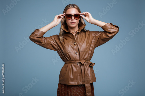Foto Fashion photo of a beautiful elegant young woman in a pretty brown leather jacket, brown long skirt, stylish sunglasses  posing over blue background