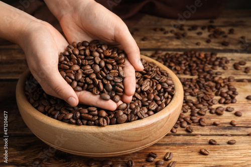 Woman taking pile of roasted coffee beans from bowl at wooden table, closeup