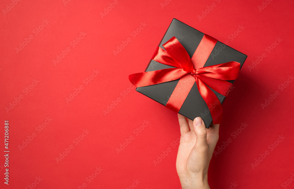 First person top view photo of girl's hand holding black giftbox with red ribbon bow on isolated red background with empty space First person top view photo of girl's hand holding black giftbox with red ribbon bow on isolated red background with empty space