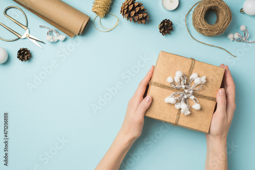 First person top view photo of hands holding kraft paper giftbox decorated with snow branch and twine white christmas tree balls handicraft tools on isolated pastel blue background with empty space
