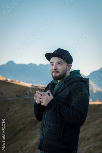 Bearded traveler man drinks tea or coffee from metal cup while resting on a rock