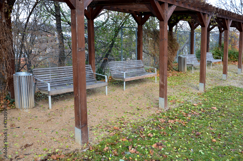 benches placed with their backs to the rope fence, in front of a wire gray galvanized fence like a cage. some are under a wooden pergola. simple metal bench with wooden planks paneling